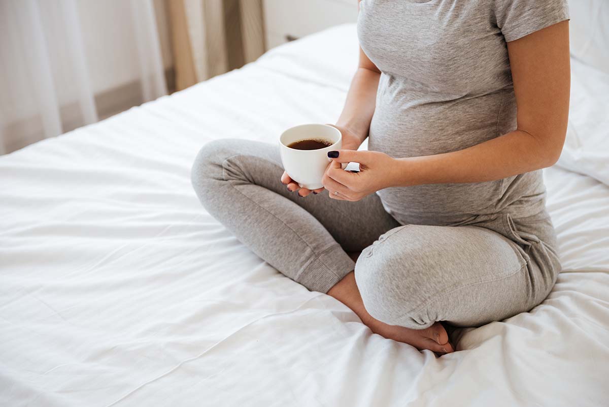 Pregnant mom in gray maternity tracksuit face not shown sits on bed holding a cup of black coffee
